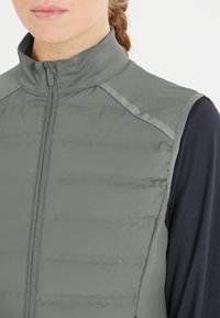 Grå zip-up väst med kviltad textur, hög krage och reflexaccenter på axlarna, parat med en mörk topp under.