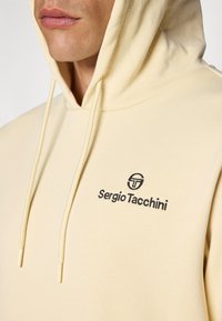 Kaki spalvos gobtuvė su raišteliu, minkšta tekstūra ir mažu juodu siuvinėtu logotipu krūtinėje su prekės ženklu Sergio Tacchini.