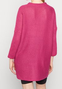 Översized, stickad tröja i stark rosa. Har ribbad struktur, rund halsringning och trekvartsärmar. Muddarna är något längre bak.