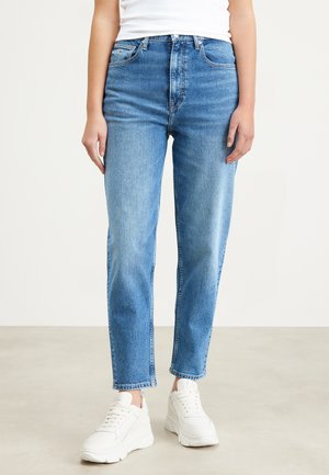 Personne portant un jean bleu clair taille haute droit, des baskets blanches et un débardeur blanc, debout sur un sol uni.