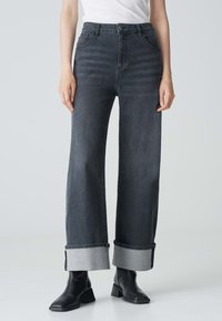 Jeans larghi grigio scuro con vita alta, orli arrotolati con tessuto a contrasto e tasche anteriori. Indossati con stivali neri alla caviglia.