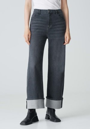 Jean large noir gris avec une taille haute, des ourlets retroussés en tissu contrastant et des poches avant. Porté avec des bottines noires.