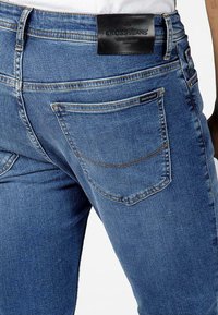 Blue denim jeans feature a slim fit, contrast stitching, and a leather label on the waistband. The back pockets are visible and lightly textured.