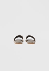 Ζευγάρι καφέ φλατ σανδάλια με μία καμπυλωτή λωρίδα και διακόσμηση από στρας γύρω από τις άκρες της σόλας, εμφανιζόμενα από πίσω.