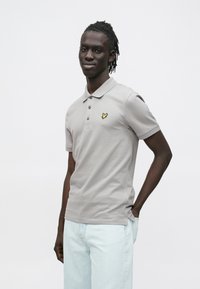 Sivá polokošeľa s golierom, tromi gombíkmi a žltým vyšívaným logom na ľavej strane hrudníka. Krátke rukávy, hladká textúra.