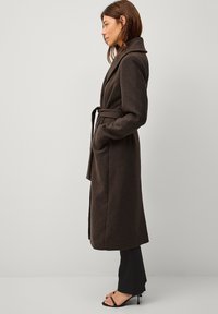 Cappotto di lana marrone con cintura, colletto a punta, tasche laterali, maniche lunghe e design a lunghezza media, abbinato a pantaloni neri e sandali con cinghie.