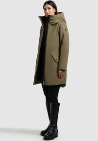 Oliwkowa kurtka puchowa z wysokim kołnierzem, zapięciem na zamek z przodu i kieszeniami na guziki. Czarne wykończenia i akcenty. Para czarnych kozaków sięgających do kolan.