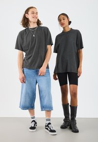 Twee modellen dragen oversized donkergrijze t-shirts. Eén draagt een lichte denim culotte en zwarte sneakers; de ander draagt een zwarte fietsbroek en hoge laarzen.