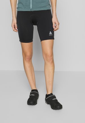 Persona con pantalones cortos de ciclismo acolchados negros y zapatos de ciclismo negros de pie contra un fondo gris.