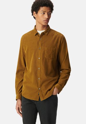 Mosterd kleurige corduroy overhemd met een button-down kraag, een enkel borstzakje, subtiele textuur en zilveren knopen, gedragen over zwarte jeans.
