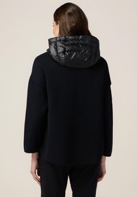 Sort strikket top med en puffy sort hætte. Designet har løse ærmer og en rund afslutning, der understreger komfort og varme.