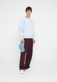 Young man wearing light blue sweatshirt and dark pants, holding a blue cap, standing against a plain white background.