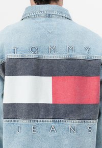 Svijetlo plavi traper jakna s dizajnom na leđima koji ima blok uzorak u tamnoplavoj, bijeloj i crvenoj boji. Ispod se nalazi izvezeni natpis "TOMMY", a ispod njega "JEANS".