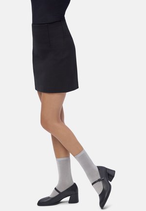 Donna che indossa una gonna nera, calze alla caviglia grigie e scarpe Mary Jane con tacco a blocco nere, in piedi con le gambe incrociate su uno sfondo bianco.
