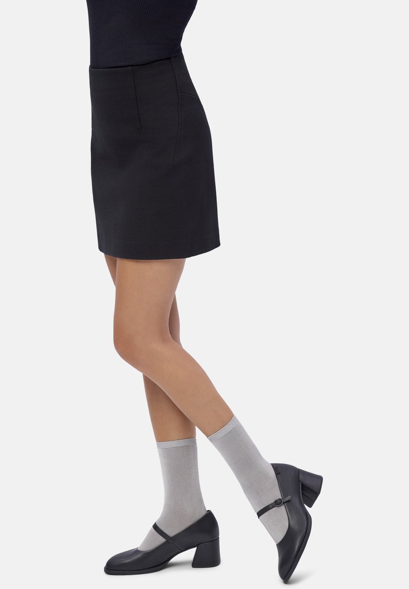 Woman wearing a black skirt, gray ankle socks, and black Mary Jane block heel shoes standing with crossed legs on a white background.