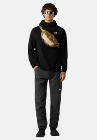 Czarna bluza z kapturem i kieszenią kangurką, zestawiona z ciemnymi spodniami i brązową torbą na ramię. Model nosi białe okulary przeciwsłoneczne i czarne obuwie.