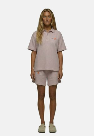 Femme aux longs cheveux blonds porte un t-shirt rose clair texturé à manches courtes et un short assorti avec des chaussures beiges à enfiler, debout sur un fond blanc.