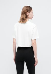 Femeie cu părul ondulat până la umeri, purtând cercei rotunzi din aur, un tricou alb scurt și pantaloni negri mulați, întorcându-se cu spatele împotriva unui fundal simplu.