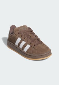 Brown suede sneaker with white three-stripe design, round toe, and textured brown laces. Features a gum rubber outsole and logo on tongue.