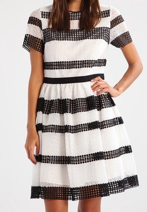 Femme portant une robe blanche mi-longue avec des bandes horizontales en dentelle noire et des manches courtes, posant avec une main sur la hanche.