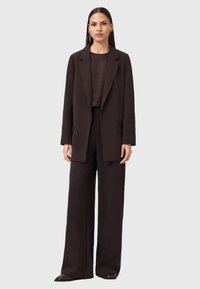 Una donna in piedi, rivolta in avanti, indossa un blazer marrone scuro, pantaloni a palazzo abbinati, un top con texture e stivali alla caviglia su uno sfondo grigio chiaro.