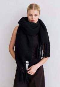 Blonde vrouw in een grote zwarte franjesjaal over een donker mouwloos topje, staand tegen een eenvoudige lichte achtergrond.