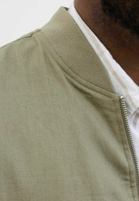 Groene bomberjack van een gestructureerde, geweven stof. Heeft een opstaande kraag en een ritsluiting, gedragen over een wit shirt.
