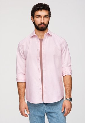 Mężczyzna z brodą w jasnoróżowej, podwiniętej koszuli na guziki, założonej na fioletowy t-shirt i jasnoniebieskie jeansy, stojący na neutralnym tle.