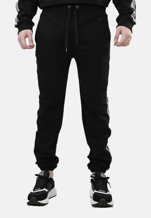 Mies, joka pukeutunut mustiin juoksuhousuihin, joissa on sivulla logoteksti, ja mustavalkoisiin lenkkareihin, seisoo kädet rentoina sivuilla.