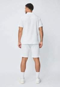 Chemise blanche à manches courtes et shorts assortis en tissu texturé, avec des rayures verticales. Porté avec des chaussettes et des chaussures blanches.
