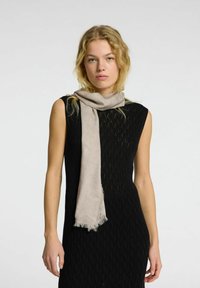 Lys beige tørklæde lavet af bl�ødt stof, med frynset kanter. Båret afslappet over en struktureret sort kjole med en tætsiddende silhuet.