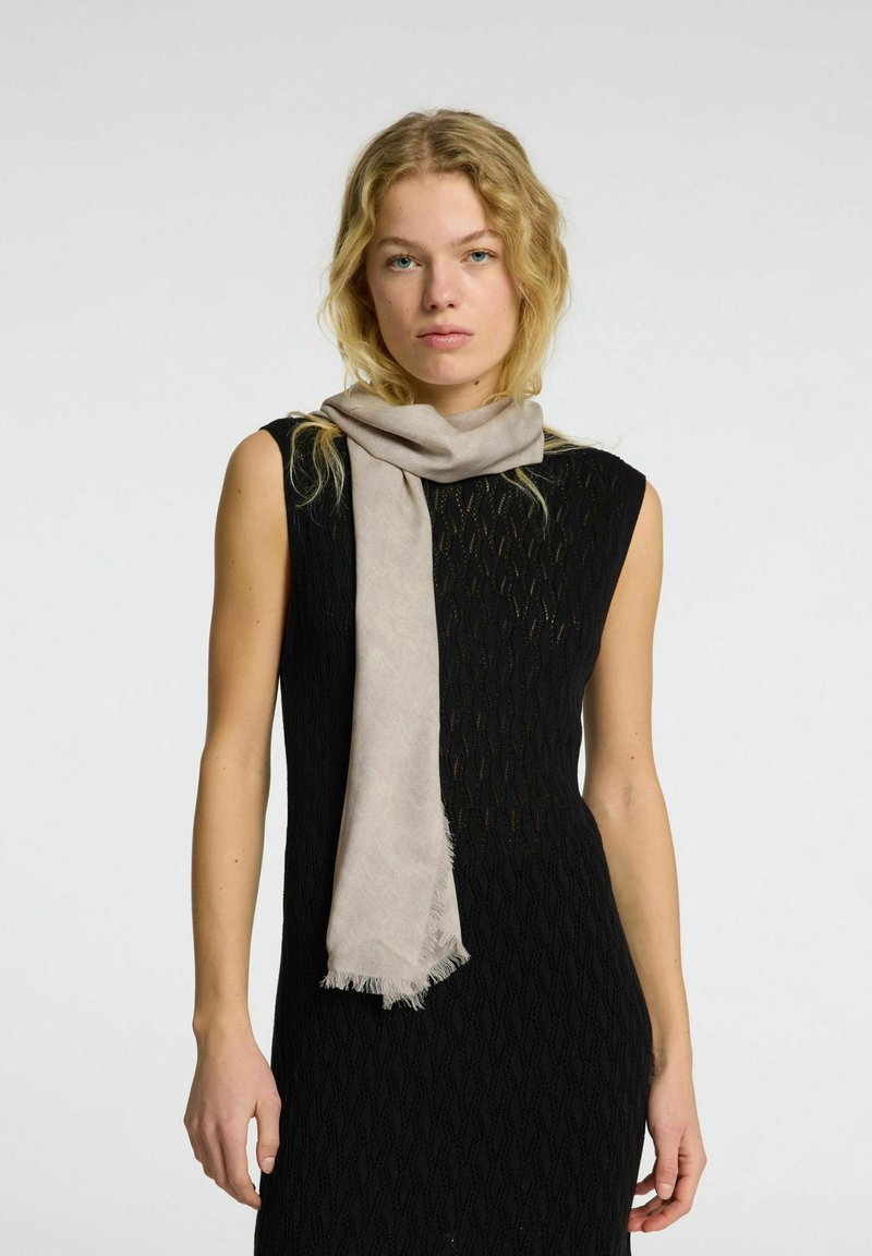 Lys beige tørklæde lavet af blødt stof, med frynset kanter. Båret afslappet over en struktureret sort kjole med en tætsiddende silhuet.