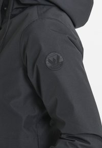 Giacca impermeabile nera con una superficie liscia, dotata di una patch rotonda nera con logo sulla parte superiore della manica e di un cappuccio.