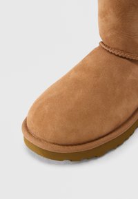 Nahaufnahme eines beigefarbenen Wildleder-Winterstiefels mit genähter Verzierung und brauner Gummisohle vor weißem Hintergrund.