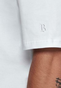 Hvid bomulds T-shirt med korte ærmer og et lille, broderet 'R' logo på venstre ærme. Glat tekstur med en klassisk pasform.