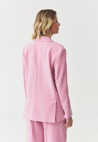 Blazer rosa con una vestibilità su misura, spacco singolo sul retro e dettagli delle cuciture minimi. Si abbina a pantaloni rosa coordinati.
