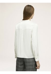 Kobieta z długimi brązowymi włosami, ubrana w białą bluzkę z długim rękawem i szare spodnie w kratę, pokazana od tyłu na neutralnym tle.