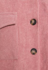 Tessuto corduroy rosa con righe verticali, dotato di una tasca sul petto e due bottoni rotondi scuri con quattro fori ciascuno.