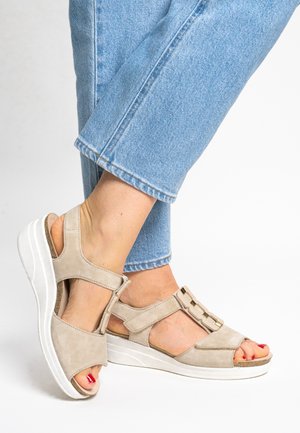 Beige suede wedge sandals with an open toe design, featuring gold accent hardware and a contrasting white sole. Paired with light denim.