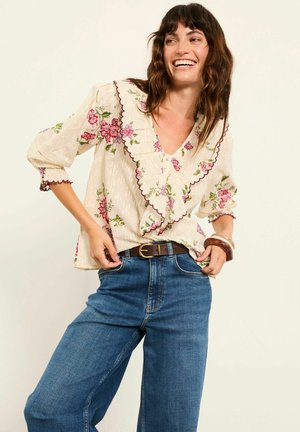Cremefarbene Bluse mit Blumenmuster, gerafftem Ausschnitt und Manschetten, kombiniert mit hoch taillierten blauen Jeans. Akzentuiert mit dunkelbraunem Gürtel. Glatte Textur.