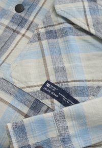 Niebieska i kremowa koszula flanelowa w kratkę, z ciemnymi guzikami i metką. Materiał prezentuje różne odcienie niebieskiego i brązowego.