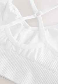 Reggiseno sportivo bianco con tessuto morbido e liscio, caratterizzato da una fascia a coste, scollo rotondo e molteplici spalline incrociate sulla schiena.