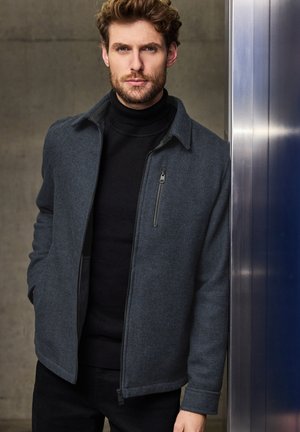 Donkergrijze geweven jas met een ritszak, kraaghals en een slim fit, gedragen over een zwarte turtleneck, tegen een neutrale achtergrond.