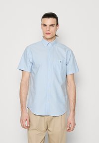 Camisa de botão de mangas curtas, na cor azul clara, com um bolso no peito e detalhe do logotipo, feita de um tecido suave com uma textura lisa.