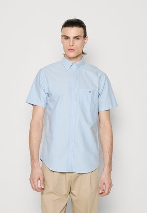 Homme aux cheveux plaqués en arrière, portant une chemise bleu clair à manches courtes boutonnée et un pantalon beige, debout devant un fond blanc uni.