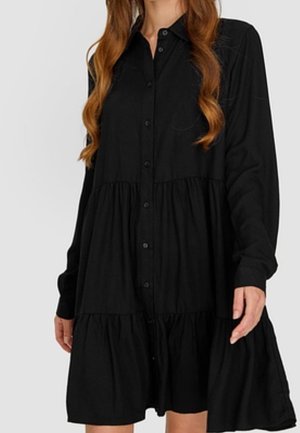 Femme portant une robe noire à manches longues et à volants, avec des boutons sur le devant, se tenant devant un fond uni.