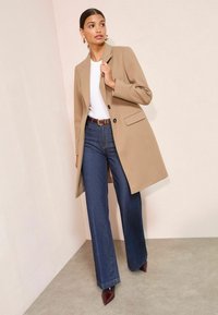 Béžový kabát so štruktúrovaným dizajnom, čiernymi gombíkmi, v kombinácii so širokými modrými džínsami a bordovými špicatými lodičkami, na pozadí svetlej steny.