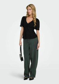 Femme portant un haut à manches courtes en dentelle noire, un pantalon ample vert foncé, des talons noirs, tenant un sac à main noir, debout sur un fond uni.