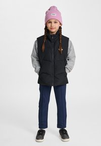 Schwarze Steppweste über einem grauen Langarmshirt, marineblaue Hose und schwarze Sneakers. Pinke Mütze mit einem weißen Etikett. Zwei Zöpfe.