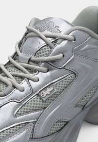 Detailný záber na strieborno-šedú tenisku Buffalo s pásmi z sieťoviny, hrubými šnúrkami a štítkami s logom na jazyku a boku.